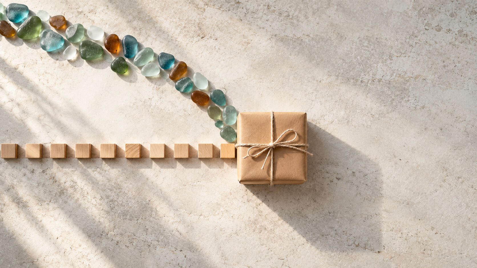Meta Ads vs Google Ads for e-commerce concept: a straight row of wooden blocks and a curving trail of colored sea-glass pebbles meeting at a kraft-wrapped parcel on a pale stone surface